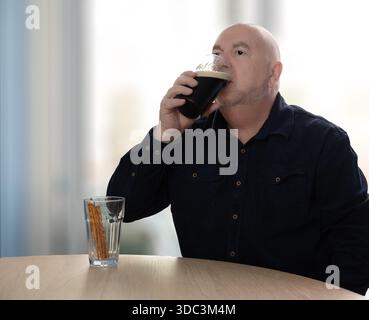A bald man in a dark blue shirt drinks a dark beer from a glass, with pretzel sticks in a separate glass on the table. New Years resolutions Stock Photo