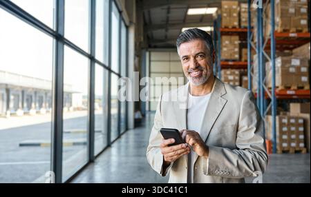 Warehouse manager standing near cardboard boxes using smartphone. Stock Photo