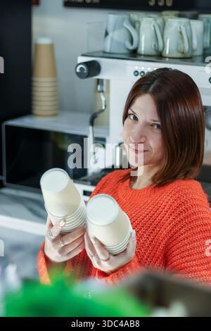 Cheerful worker in vibrant orange knitwear showcasing disposable cups in cafe equipped with coffee maker and assorted mugs. Stock Photo