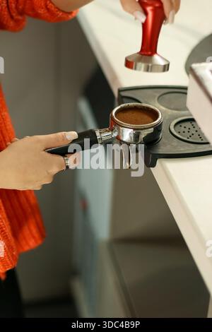 Close-up of female barista's hands tamping fresh ground coffee into a metal portafilter on a counter. Stock Photo