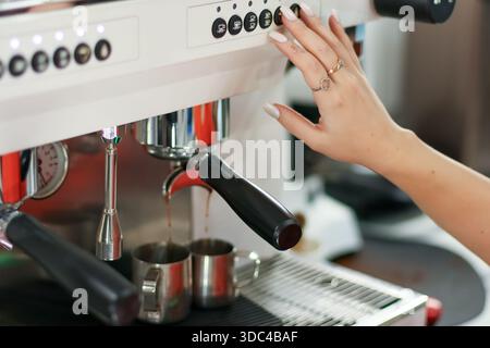 Woman's manicured hand activating espresso machine, dark coffee streaming into two metal mugs on black mat Stock Photo