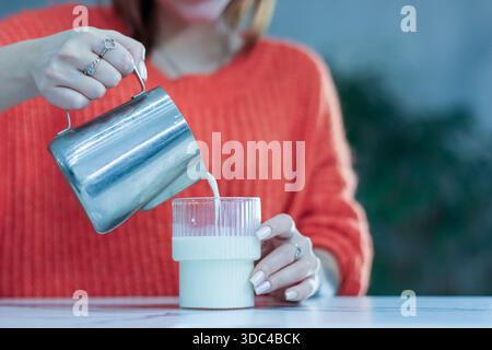 Extreme close-up of hands pouring white frothed milk from stainless steel pitcher into glass cup on white marble surface Stock Photo