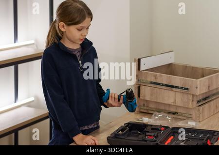 Preteen girl in navy fleece holding blue cordless drill with both hands, preparing to work on wooden drawer unit Stock Photo