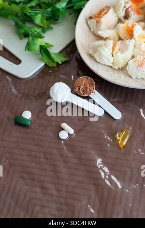 Close-up of halved boiled eggs on plate, fresh parsley, tomato slices, protein powders on spoons, and scattered vitamin pills on plastic-covered table Stock Photo
