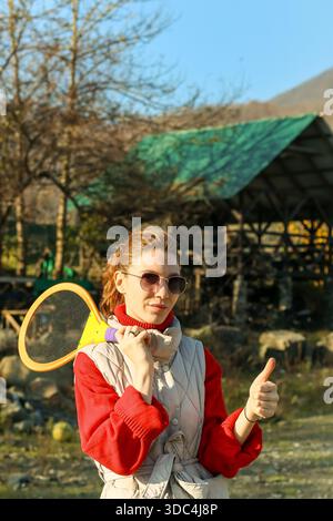 Young woman in sunglasses smiles with thumbs up, racket over shoulder, standing in mountain park with green pavilion and trees. Stock Photo