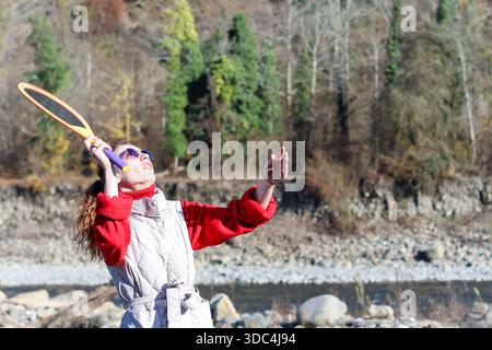 Young woman in casual attire raises colorful badminton racket high, focusing upward near rocky riverbank with forested background. Stock Photo