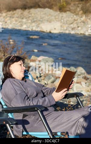 A woman in gray attire holds an open book while resting hand on chin, seated on chair facing a flowing river with stony shores. Stock Photo