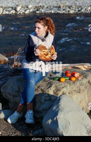 Woman in jeans and vest sits on riverbank rocks, firmly holding a stuffed flatbread as tomatoes, cucumbers, and oranges rest on a box beside her. Stock Photo