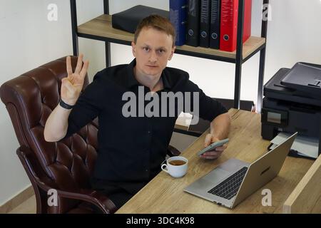 Professional at workstation shows three-finger gesture with one hand while using mobile device beside computer and tea. Stock Photo