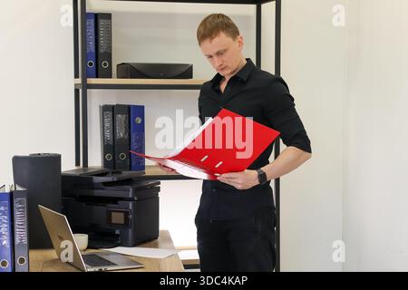Business attire male stands holding and reading from open red folder beside desk in clean office interior. Stock Photo