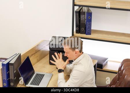 Male in office raises hand with fingers splayed toward device on desk, accompanied by laptop, files, and cup in bright professional space. Stock Photo