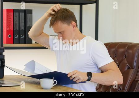 Young adult male in white shirt sits at wooden desk, puzzled expression, hand on head, open blue folder with papers, cup of beverage nearby, office sh Stock Photo