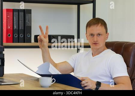 Casual office worker gesturing peace sign with documents on desk and coffee nearby Stock Photo