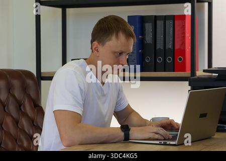 Focused young adult male working on silver laptop at wooden desk in modern office space with colorful file folders and printer in background. Stock Photo