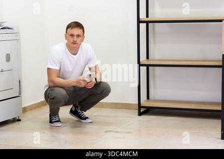 Man squatting and staring seriously at phone in empty room. Adult man crouching on tiled floor in minimal room with empty shelves, holding and gazing Stock Photo