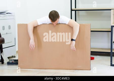 Exhausted man resting arms and head on cardboard sheet. Tired young man leaning on big cardboard panel with arms draped over, showing fatigue during D Stock Photo