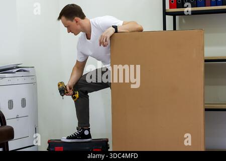 Man standing on toolbox drilling into large cardboard box in office. Casual worker balances on tool case, using power drill on side of tall brown card Stock Photo