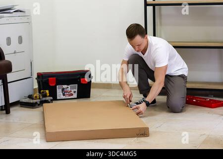 Man carefully cutting cardboard with knife on the ground. Young adult male kneeling and using cutter to slice through cardboard material during DIY pr Stock Photo