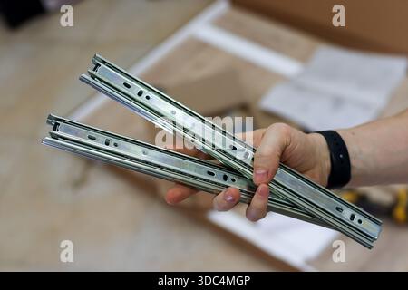 Close-up of hand holding drawer runner hardware. Hand presenting pair of metallic telescopic drawer slides against blurred background of furniture par Stock Photo