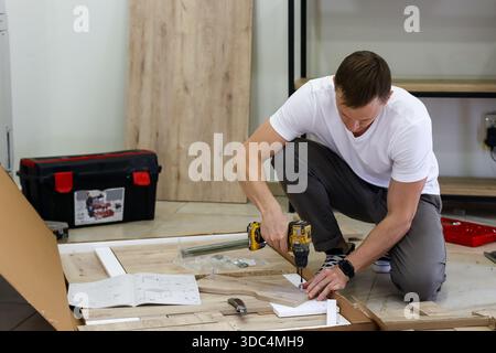 Focused man assembling flat-pack furniture with power drill. Man in casual clothes kneeling and using yellow drill on wooden panels surrounded by asse Stock Photo