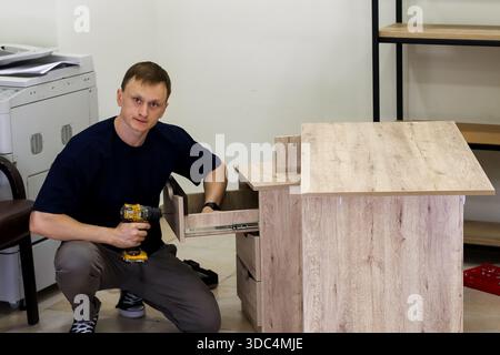 Man installing drawer slides inside wooden cabinet. Close view of worker using cordless drill to mount metal slides on drawer in light wood furniture Stock Photo
