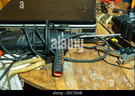 Power drill with a coiled cable and a strong drill bit laying on a rustic wooden workbench. Surrounded by various hand tools like pliers and a staple Stock Photo