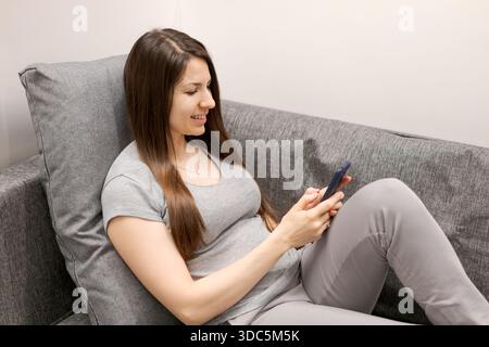 A woman is sitting on a couch and looking at her cell phone. She is smiling and she is enjoying herself Stock Photo