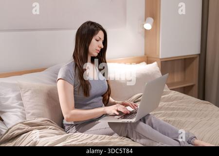 A woman is sitting on a bed with a laptop in front of her. She is typing on the laptop and she is focused on her work. Concept of productivity and con Stock Photo