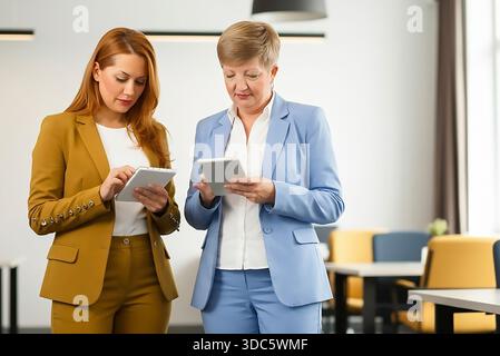 Two women sitting on a couch, both using laptops. One of the women is wearing a yellow jacket Stock Photo