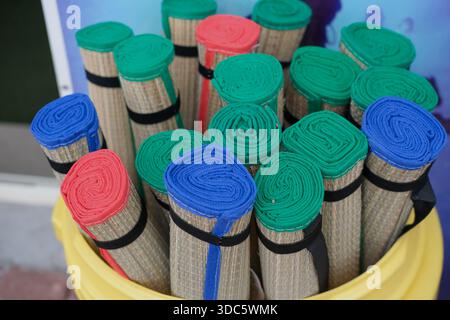 Top-down view of rolled straw mats bound with black straps, green blue and red fabric edges visible, placed in a yellow plastic container. Stock Photo