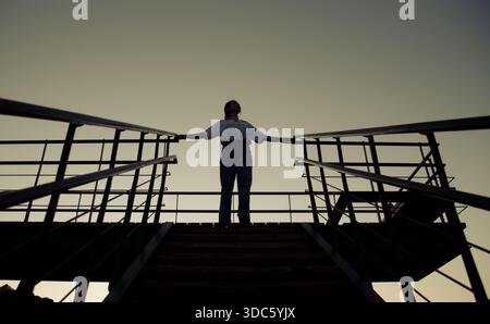 A woman stands on a high platform with her arms outstretched as she gazes at the horizon during sunset. The sky has a mix of colors as the day ends. Stock Photo