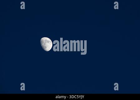 Half moon hangs in the blue sky during the late afternoon. It is bright and clearly visible against the backdrop of the sky. The surroundings are calm Stock Photo