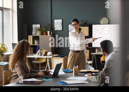 Caucasian young woman presenting data on screen to diverse colleagues in modern office, gesturing toward chart while explaining business statistics during meeting Stock Photo