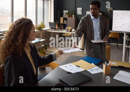 Black man handing business documents to young adult Caucasian woman during office meeting, both focused on paperwork with charts and graphs visible on table and whiteboard Stock Photo