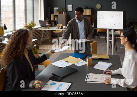 Multiethnic group of young adult professionals collaborating in modern office, Black man standing presenting documents to Caucasian woman while another woman taking notes at table Stock Photo