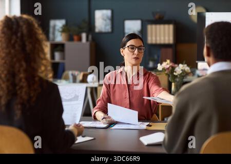 Caucasian woman handing document to Black man while sitting at desk with multiethnic colleagues during business meeting, discussing paperwork and collaborating on project Stock Photo