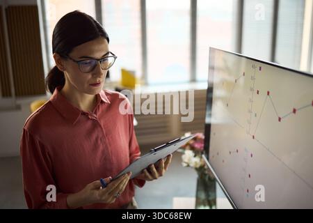 Caucasian young adult woman analyzing financial data on clipboard while standing near large monitor displaying line graphs in modern office setting, wearing eyeglasses and focused expression Stock Photo