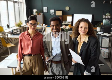 Portrait of young adult Caucasian women and young adult Black man standing together in modern office holding documents and smiling at camera Stock Photo