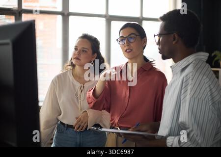 Caucasian young adult woman explaining project details to Caucasian young adult woman and Black young adult man standing together in office, collaborating while looking at computer monitor Stock Photo