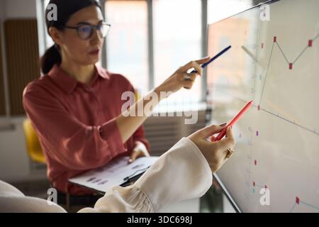 Young adult Caucasian woman explaining data on digital screen while young another woman holding clipboard analyzing graph, both collaborating on business presentation Stock Photo