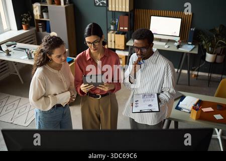 Three young adult colleagues, including Caucasian women, Black man, standing in modern office collaborating on digital tablet and analyzing documents together Stock Photo
