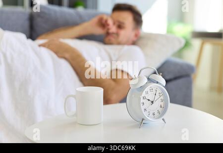 Sleepy man lying near alarm clock in bed, morning wake-up with coffee mug Stock Photo