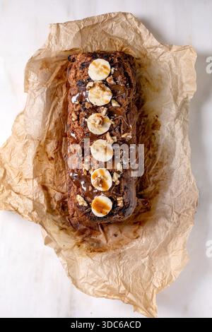 Whole homemade banana bread cake with walnuts, drizzled with caramel and topped with fresh banana. Placed on crinkled baking paper on white marble tab Stock Photo