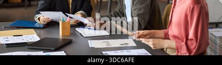 Diverse group of professionals collaborating at table, reviewing business documents and analyzing printed charts, hands of Caucasian women and Black man visible, header Stock Photo