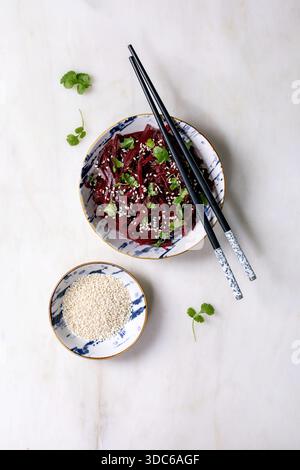 Korean-style vegan beet salad. Marinated grated beetroot with sesame seeds and coriander leaves in ceramic bowl. Nearby bowl with sesame seeds and cho Stock Photo