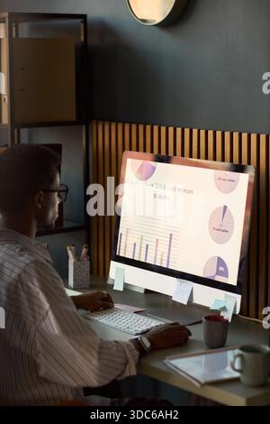 Young adult Black man working at desk analyzing financial data on large desktop computer monitor displaying charts and graphs in modern office setting Stock Photo