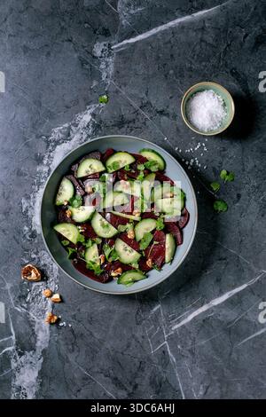 Vegan beet salad. Marinated sliced beetroot with fresh cucumber, walnuts, and coriander leaves in ceramic bowl. Nearby bowl with sea salt on black mar Stock Photo