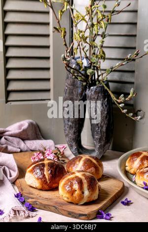 Hot cross buns on wooden cutting board with spring decoration. Easter pastry styled with willow branches in rustic vase, pink blossoms and violet flow Stock Photo