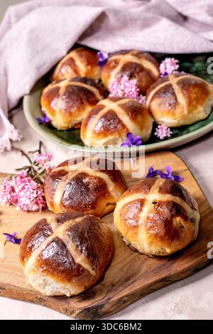 Traditional hot cross buns on wooden board. Easter baking with spices and dried fruits decorated with spring flowers. Homemade sweet bread rolls with Stock Photo