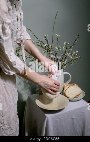 Woman linen clothing squeezes homemade cottage cheese arranging white cherry blossoms ceramic pitcher rustic table setting slice bread plate minimalis Stock Photo